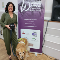 Photo of Sarah at the awards standing in front of a sign that says Inspirational Women of Sheffield. She is holding her award and her guide dog is with her
